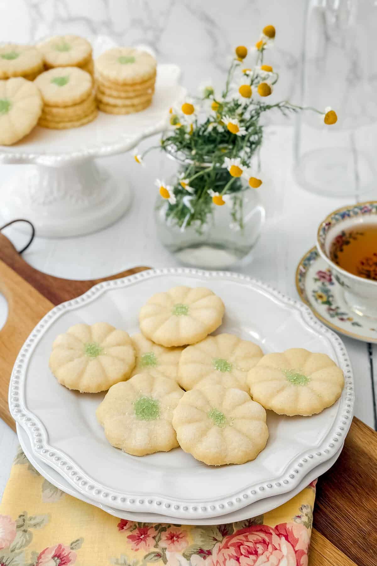 A white plate with six flower-shaped sugar cookies, reminiscent of classic Girl Scout Cookies, each with green sugar in the center, sits on a floral napkin. In the background are more cookies, a teacup with tea, and a small vase of chamomile flowers.