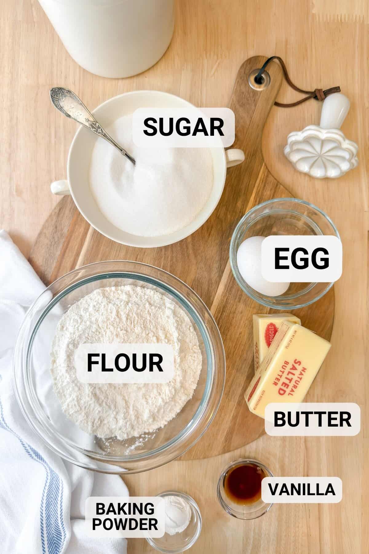 A top-down view of baking ingredients on a wooden board, perfect for making Girl Scout Cookies: a bowl of sugar with a spoon, an egg, flour, butter, baking powder, and vanilla extract.