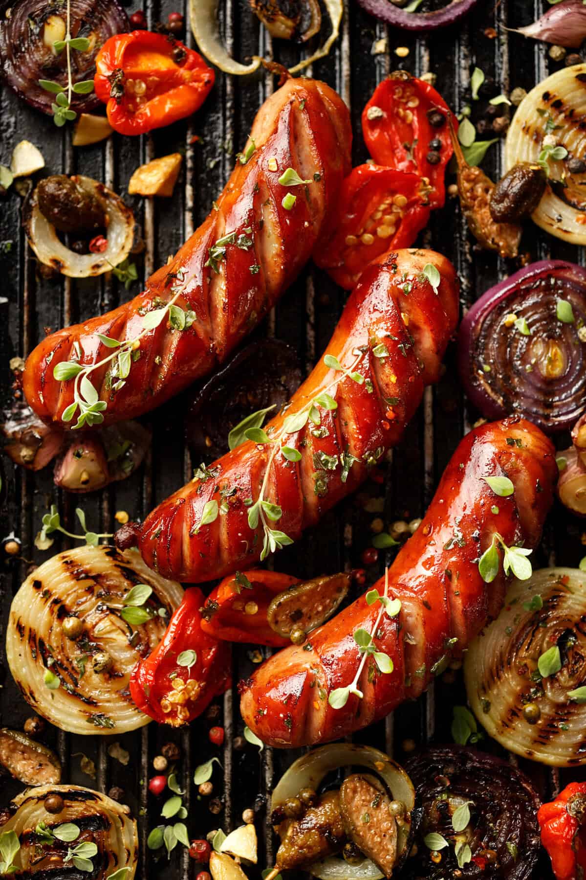 Three grilled sausages with char marks on a grill, surrounded by grilled onion slices, tomatoes, mushrooms, garlic, herbs, and peppercorns. The sausages are garnished with fresh microgreens.