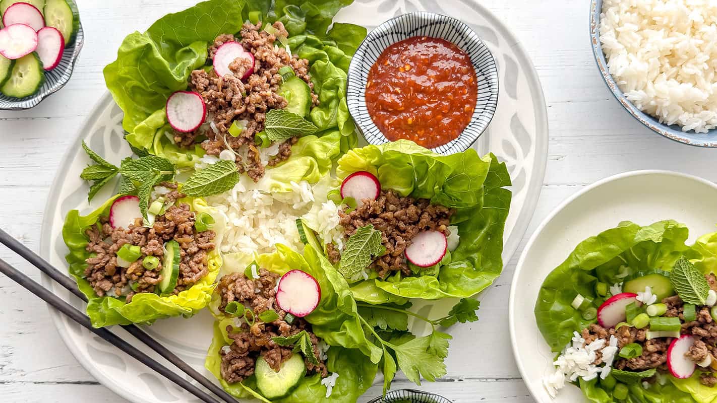 A plate of Korean beef lettuce wraps filled with ground meat, rice, cucumber, radish, and mint, served alongside a bowl of sauce and a side of white rice.