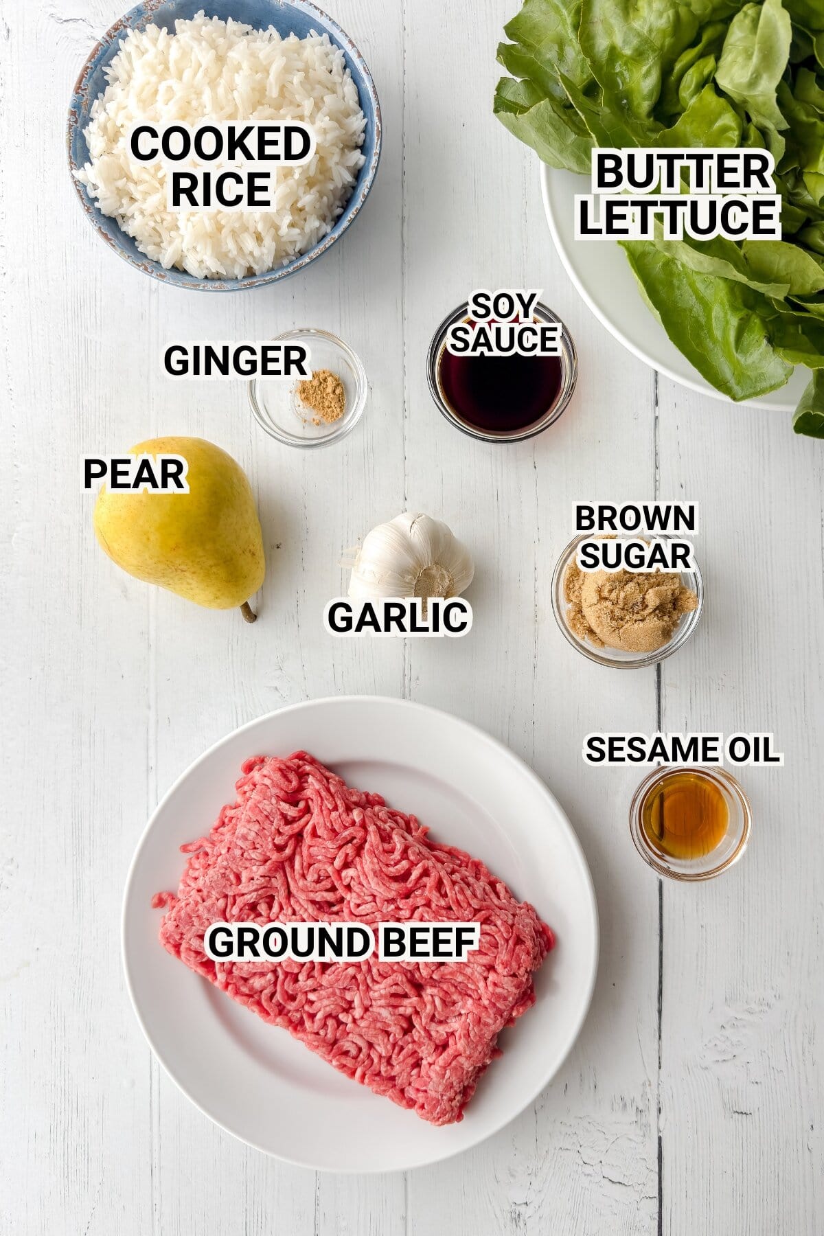 Top-down view of ingredients on a white table: cooked rice, butter lettuce, ground beef, a pear, garlic, ginger, soy sauce, brown sugar, and sesame oil, each labeled with text.