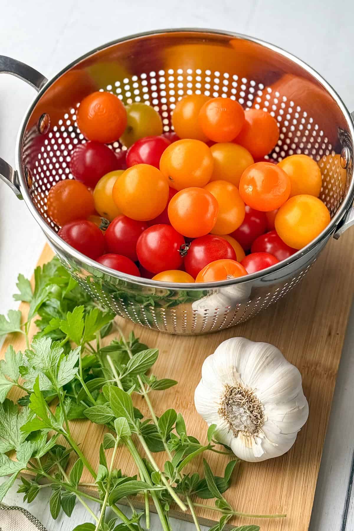 A metal colander filled with colorful cherry tomatoes sits on a wooden board next to fresh green herbs and a whole garlic bulb.