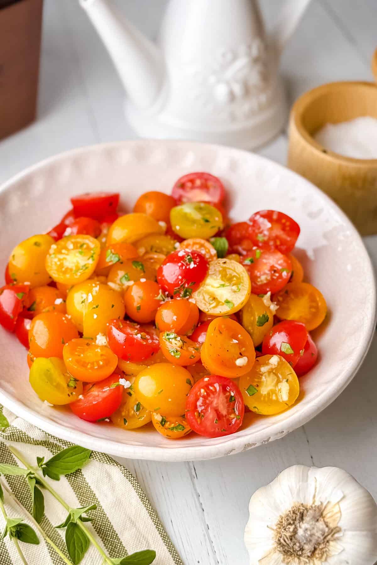 A white bowl filled with colorful halved cherry tomatoes, garnished with chopped herbs and salt. Fresh garlic, a small bowl of salt, and a teapot are nearby on a light wooden table.