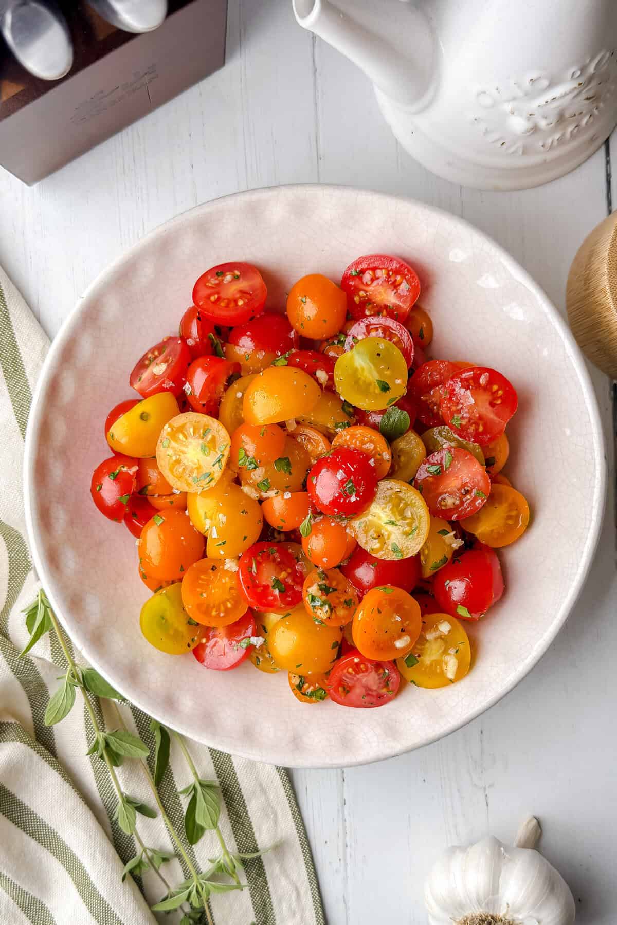 A bowl of colorful cherry tomato salad with chopped herbs on a white table, surrounded by a striped napkin, garlic, and a white ceramic jug.