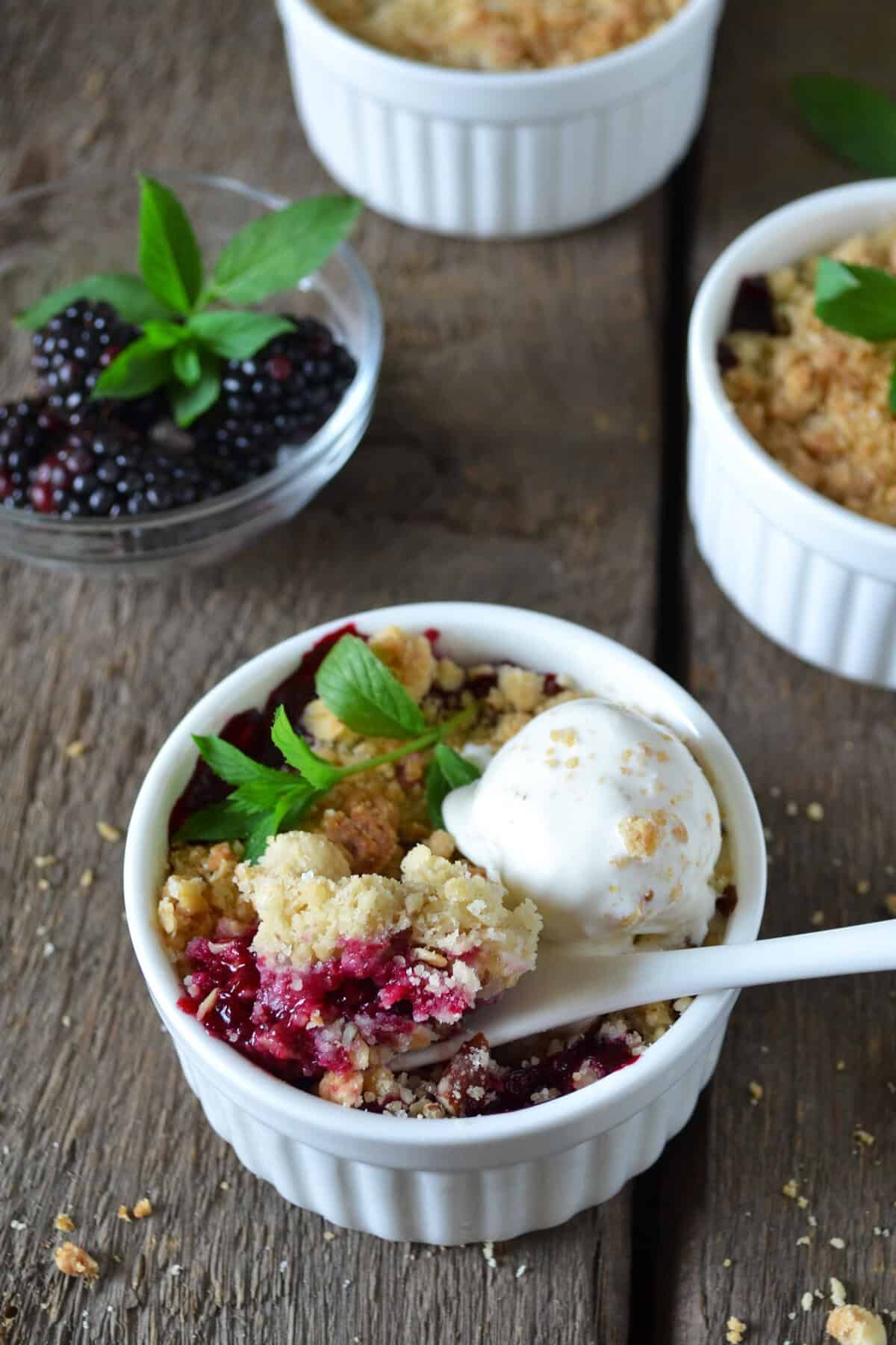 A white ramekin filled with blackberry crumble topped with a scoop of vanilla ice cream and a mint sprig, with a spoon inside. In the background, more crumbles and a bowl of fresh blackberries with mint are visible.