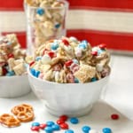 A white bowl filled with 4th of July Chex Mix featuring Chex cereal, red, white, and blue candy-coated chocolates, pretzels, and a white chocolate drizzle.