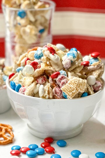 A white bowl filled with 4th of July Chex Mix featuring Chex cereal, red, white, and blue candy-coated chocolates, pretzels, and a white chocolate drizzle.