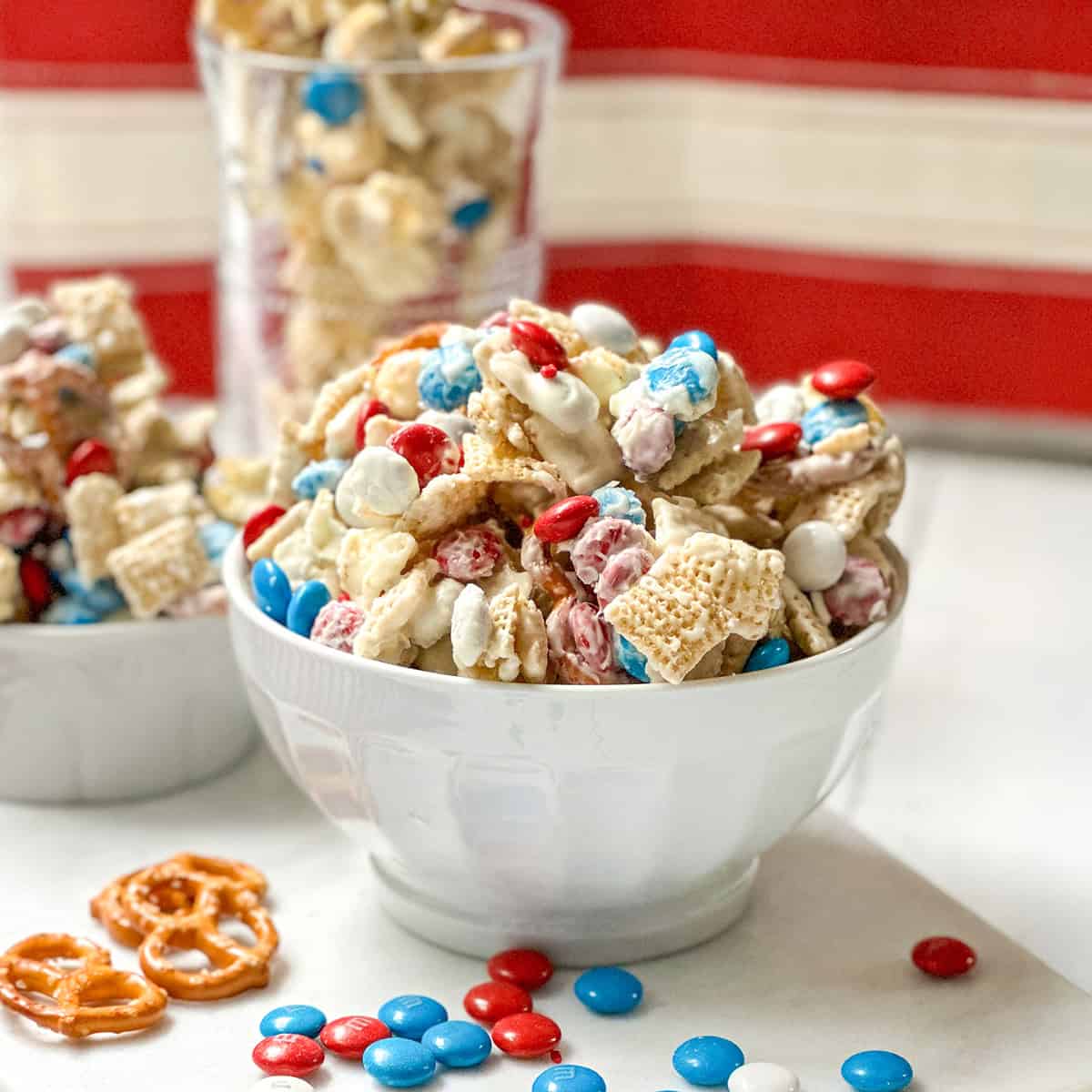 A white bowl filled with 4th of July Chex Mix featuring Chex cereal, red, white, and blue candy-coated chocolates, pretzels, and a white chocolate drizzle.