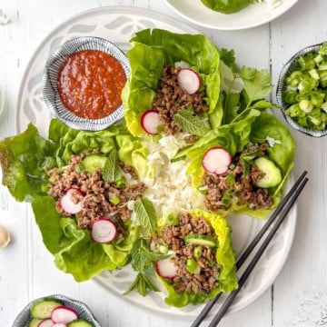 Plate of Korean beef lettuce wraps filled with ground meat, sliced radish, avocado, green onions, and mint, served with a mound of white rice, a bowl of red sauce, and a pair of black chopsticks on the side.