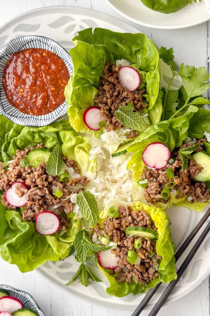 Plate of Korean beef lettuce wraps filled with ground meat, sliced radish, avocado, green onions, and mint, served with a mound of white rice, a bowl of red sauce, and a pair of black chopsticks on the side.