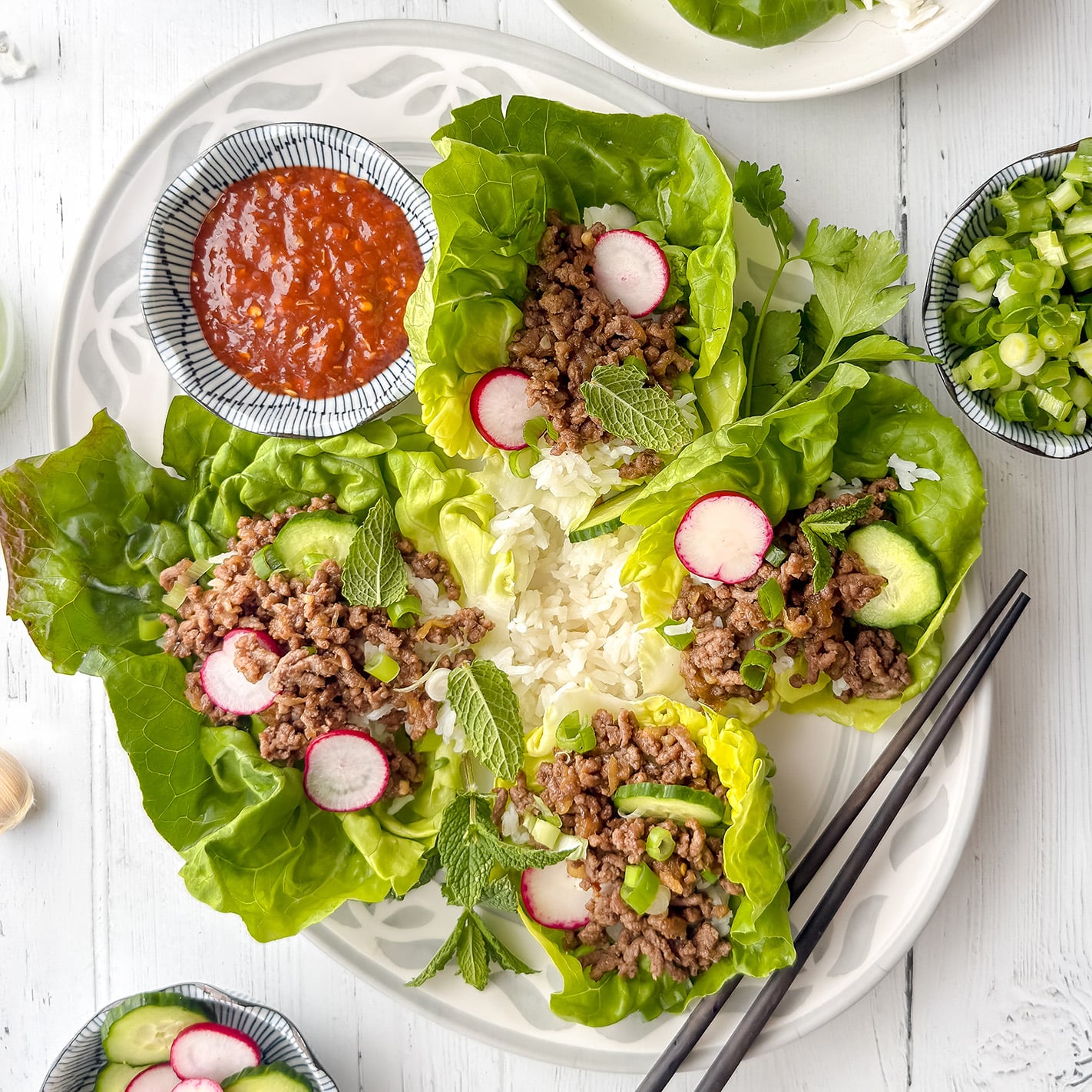 Plate of Korean beef lettuce wraps filled with ground meat, sliced radish, avocado, green onions, and mint, served with a mound of white rice, a bowl of red sauce, and a pair of black chopsticks on the side.