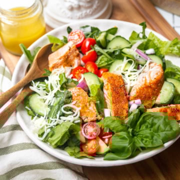 A bowl of salmon salad with grilled chicken slices, lettuce, spinach, cucumber, cherry tomatoes, red onion, and shredded cheese, served with wooden utensils and a jar of dressing on the side.