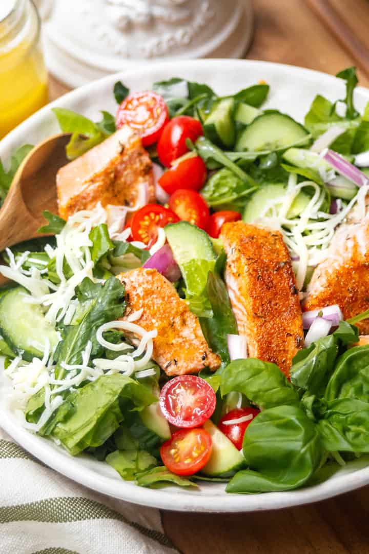 A bowl of salmon salad with grilled chicken slices, lettuce, spinach, cucumber, cherry tomatoes, red onion, and shredded cheese, served with wooden utensils and a jar of dressing on the side.