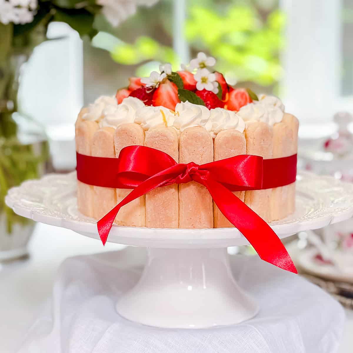 A charlotte cake decorated with ladyfingers, a red ribbon, whipped cream, strawberries, and small white flowers on a white cake stand-perfect for a delightful summer afternoon tea.