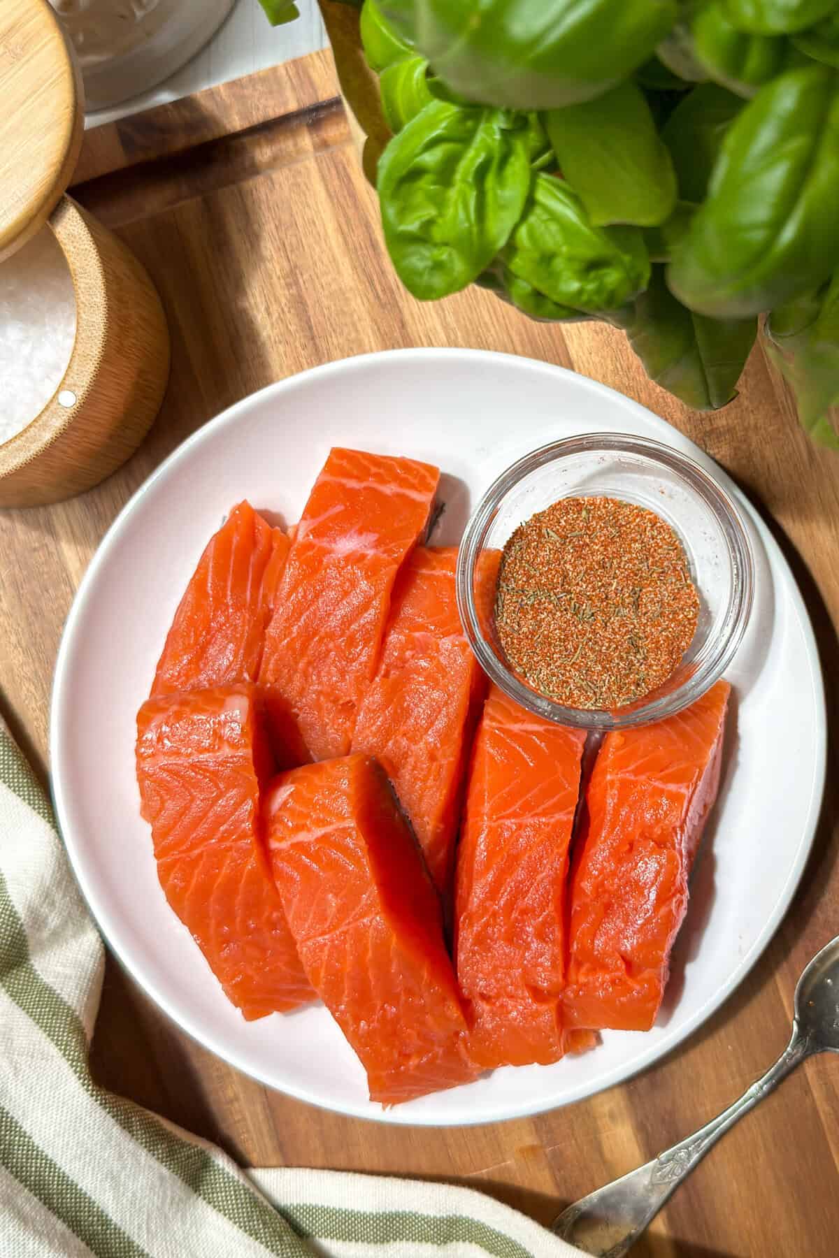 A white plate with six raw salmon fillets and a small glass bowl of seasoning sits on a wooden surface next to fresh basil, salt, and a striped kitchen towel-perfect ingredients for crafting a vibrant salmon salad.