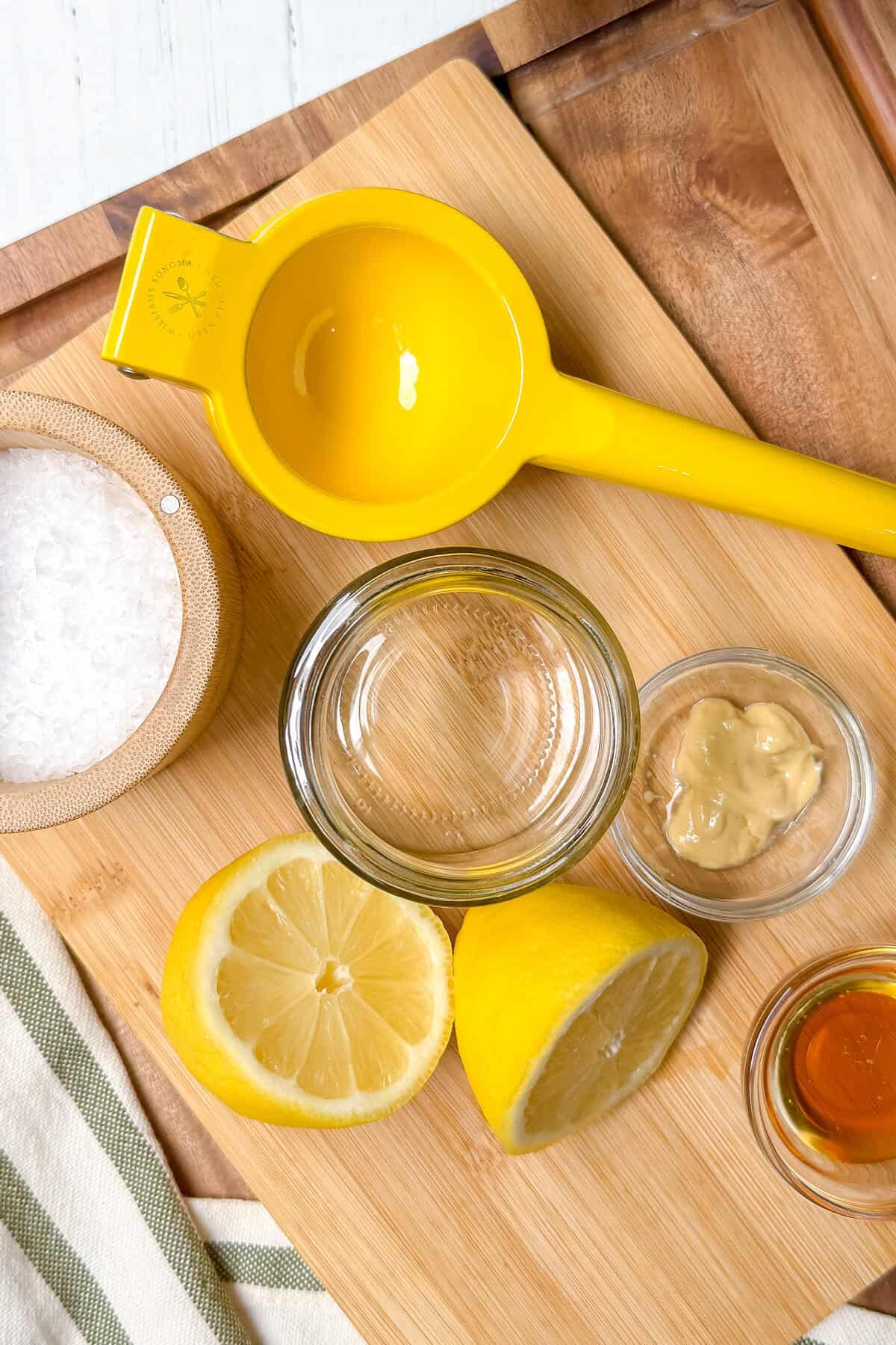 A yellow lemon squeezer, a bowl of salt, a halved lemon, an empty glass jar, Dijon mustard, and honey on a wooden cutting board-everything you need for a zesty dressing perfect for salmon salad.