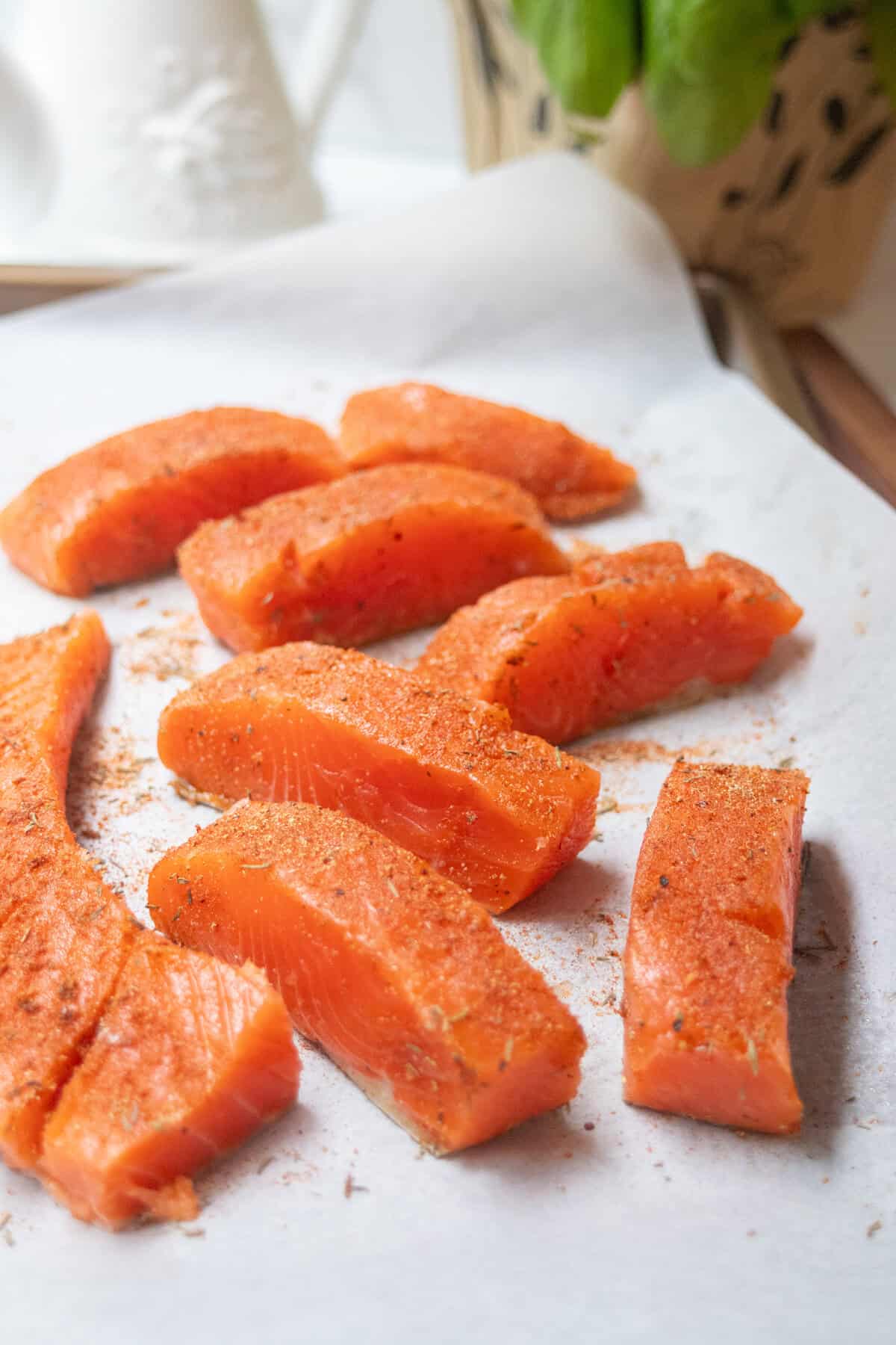 Slices of raw salmon fillet seasoned with spices are arranged on a sheet of parchment paper, ready for cooking or preparing a fresh salmon salad. A potted plant and white kitchen items are blurred in the background.