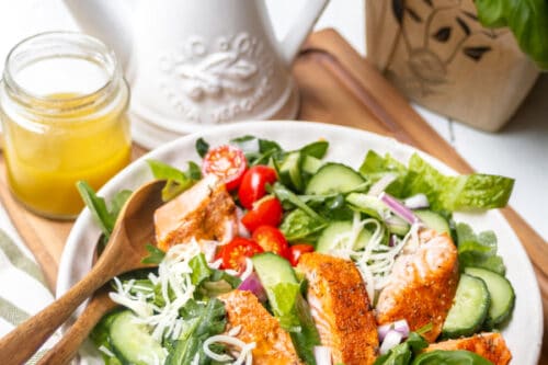 A fresh salad with grilled chicken or salmon salad, cherry tomatoes, cucumbers, spinach, shredded cheese, and red onion in a white bowl, with a fork and dressing jar beside it. Basil and an oil dispenser are in the background.