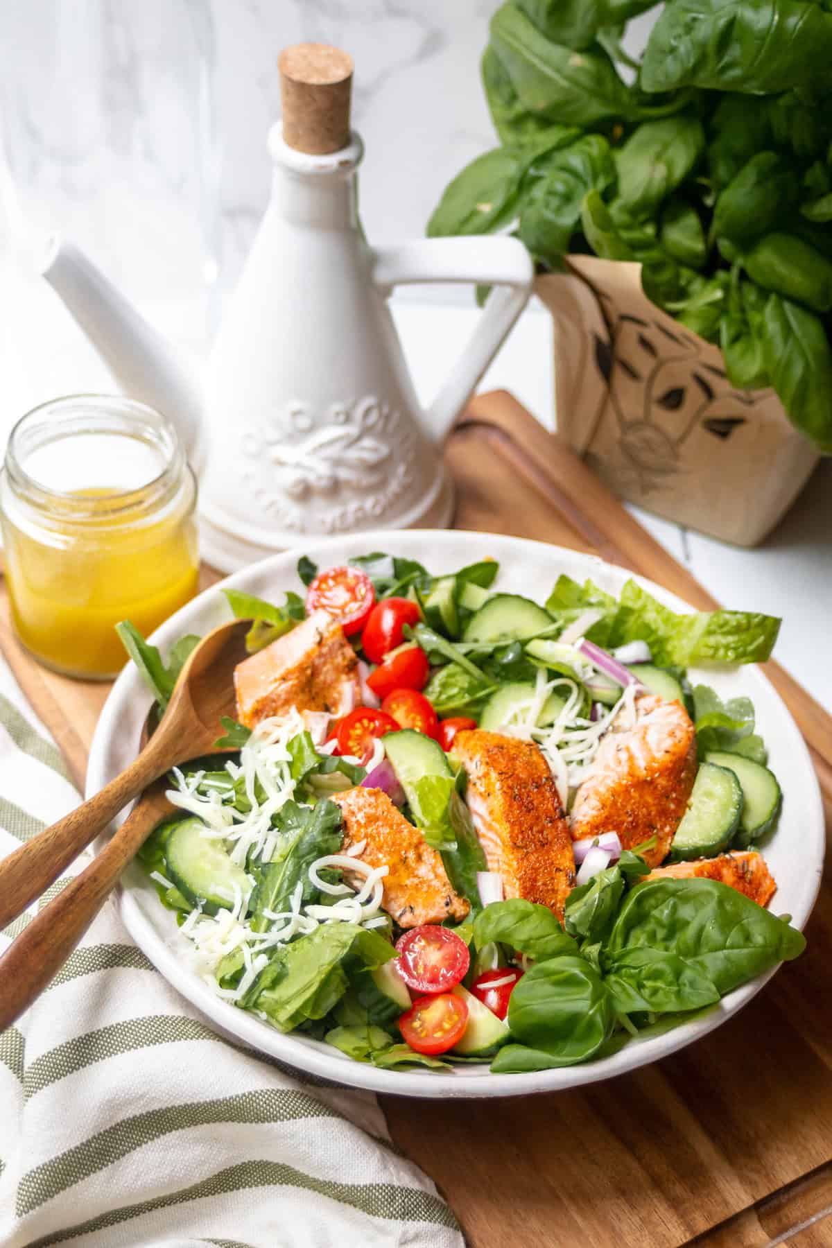 A fresh salad with grilled chicken or salmon salad, cherry tomatoes, cucumbers, spinach, shredded cheese, and red onion in a white bowl, with a fork and dressing jar beside it. Basil and an oil dispenser are in the background.