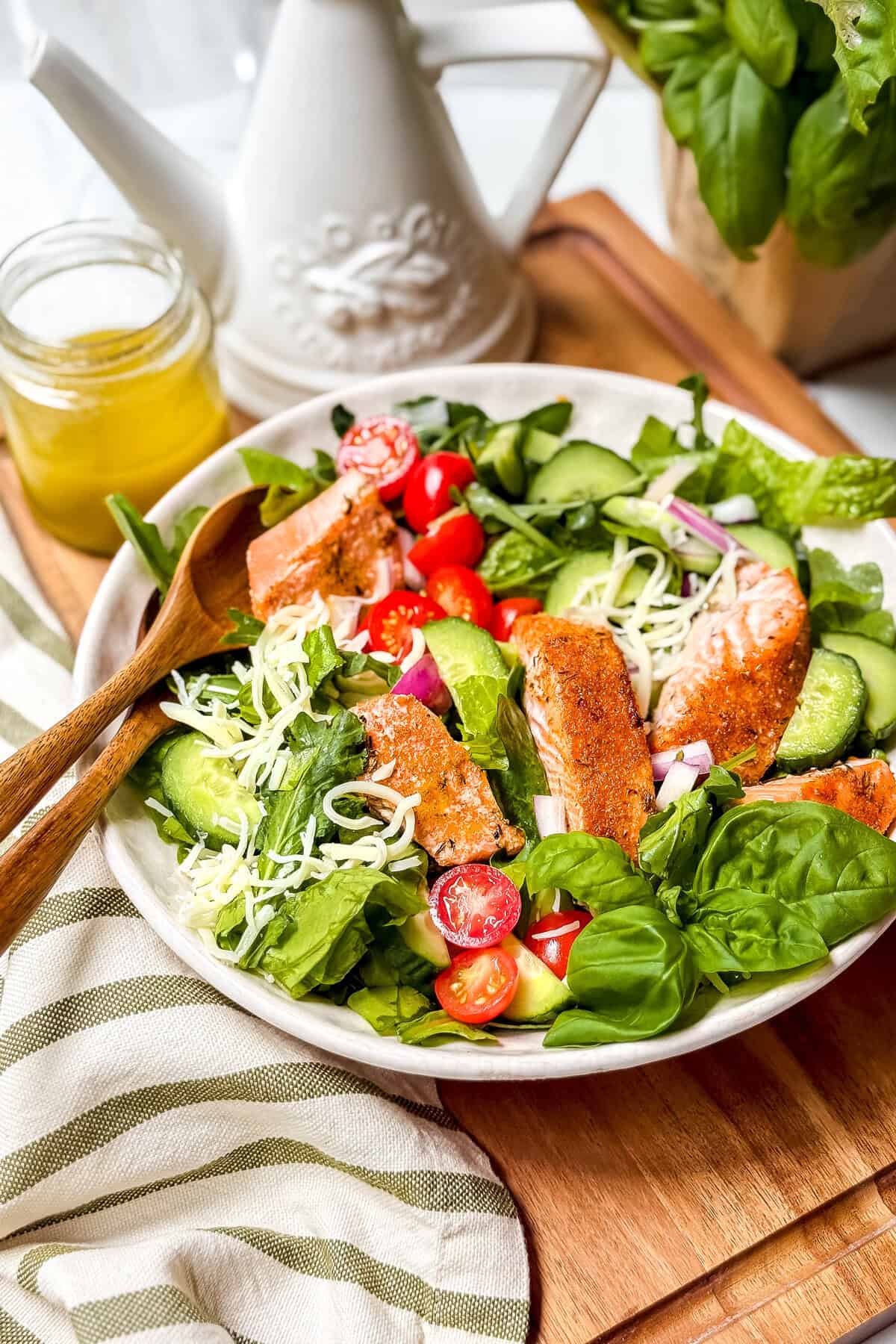 A fresh salmon salad with grilled salmon, cherry tomatoes, cucumber, shredded cheese, spinach, and mixed greens, served in a bowl with a wooden fork, next to a jar of dressing and a white oil dispenser.
