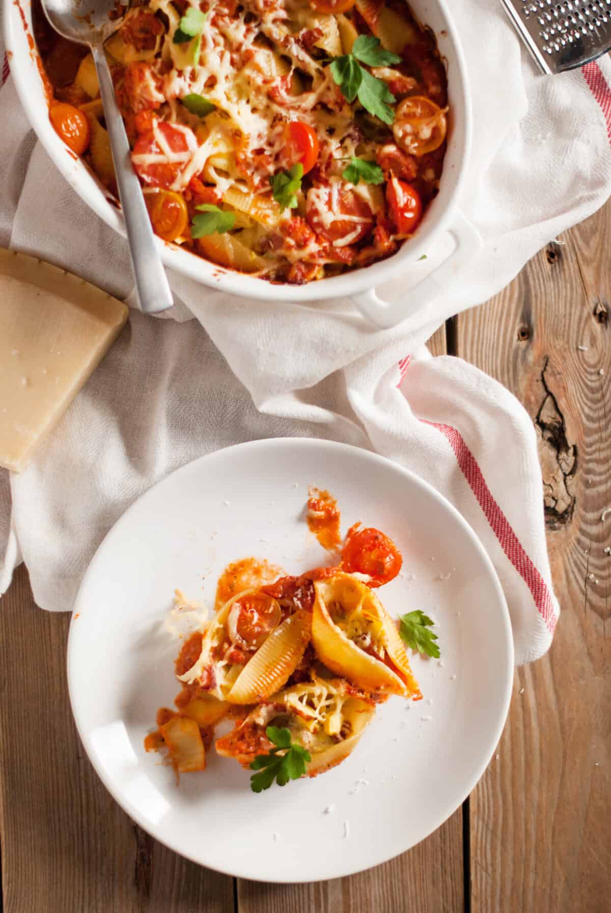 A white plate with stuffed pasta shells topped with melted cheese, tomato sauce, cherry tomatoes, and parsley sits on a rustic wooden table beside a baking dish of the same pasta, a block of cheese, and a white towel.