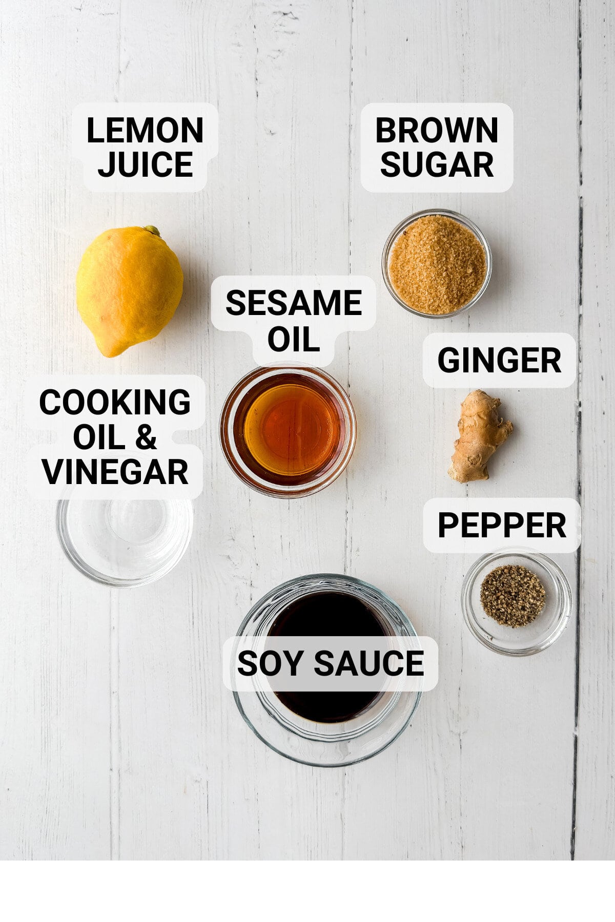 Overhead view of labeled ingredients for Asian Coleslaw on a white surface: lemon juice, brown sugar, cooking oil & vinegar, sesame oil, fresh ginger, pepper, and soy sauce.