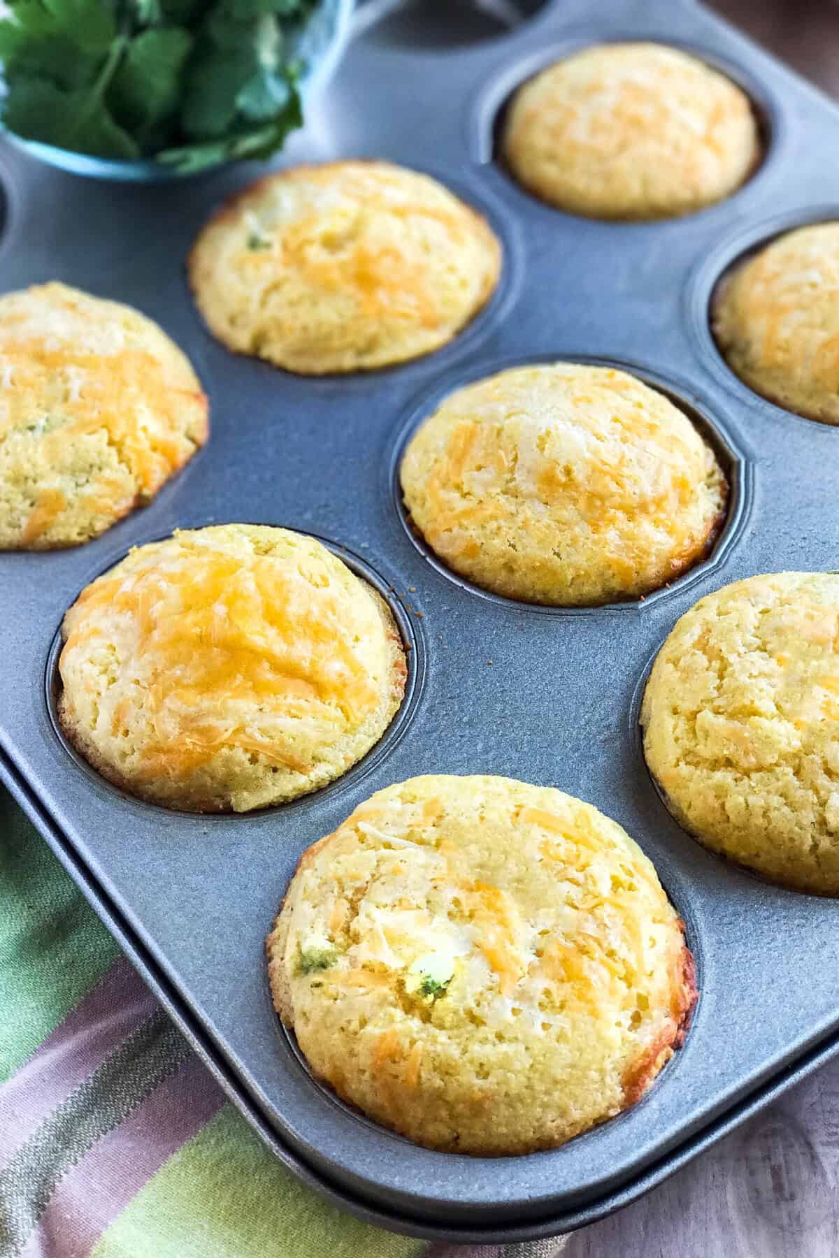 A muffin tin filled with freshly baked cheesy cornbread muffins, golden brown on top, sits on a striped cloth. A bowl of green herbs is visible in the background.