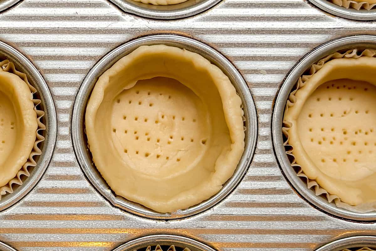 Close-up of a muffin tin with unbaked pie crusts pressed into the cups. The crusts have fork holes pricked in the bottom, ready for baking.