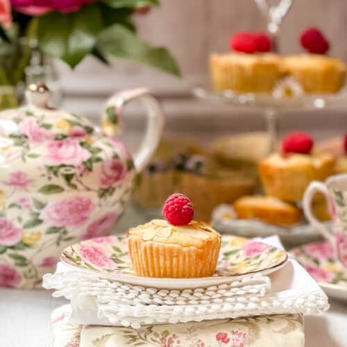 A floral teapot, teacup, and plate hold tea and a bakewell tart topped with a raspberry. More are displayed on a glass tiered stand in the background, with chamomile tea flowers and a white napkin accenting the scene.