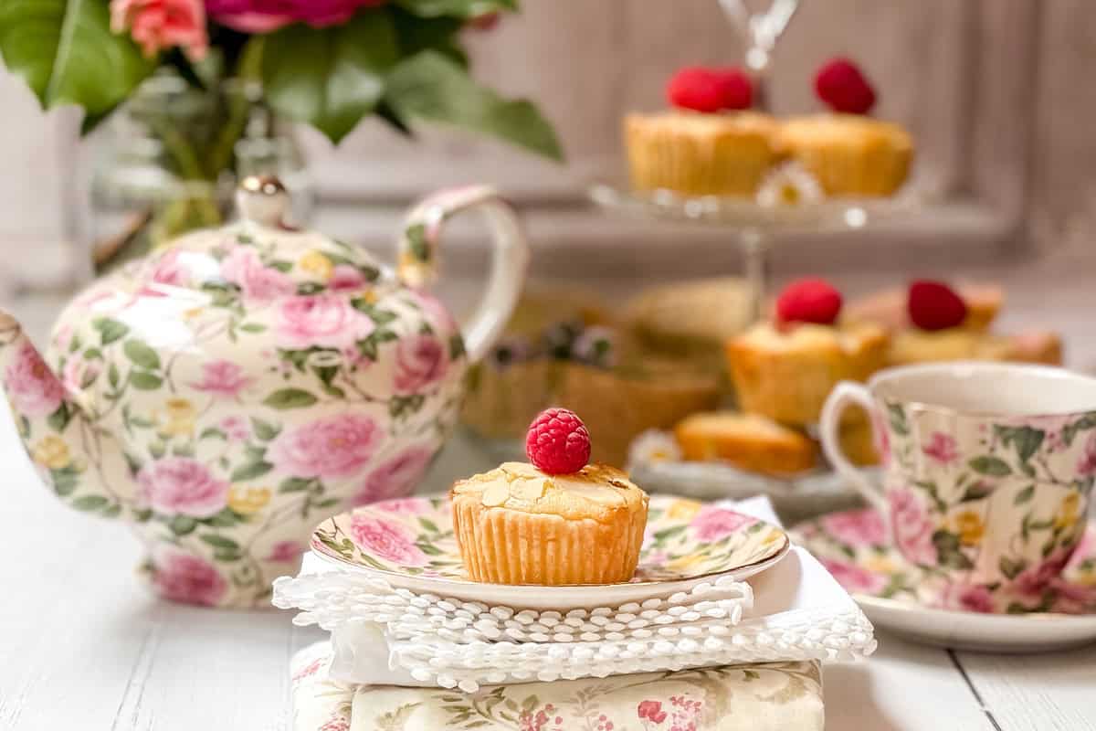 A floral teapot, teacup, and plate hold tea and a bakewell tart topped with a raspberry. More are displayed on a glass tiered stand in the background, with chamomile tea flowers and a white napkin accenting the scene.