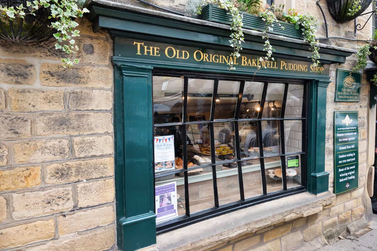 A stone building with green trim houses "The Old Original Bakewell Pudding Shop." Through the large window, pastries and cakes are visible inside. Hanging plants decorate the exterior above the shop sign.