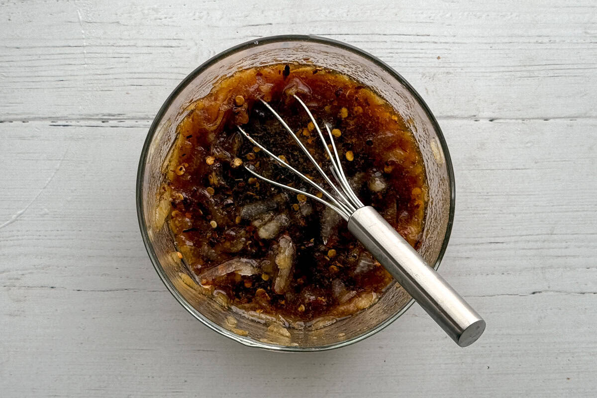 A glass bowl containing a dark, chunky marinade with visible spices and seeds, being mixed with a metal whisk, sits on a white wooden surface.