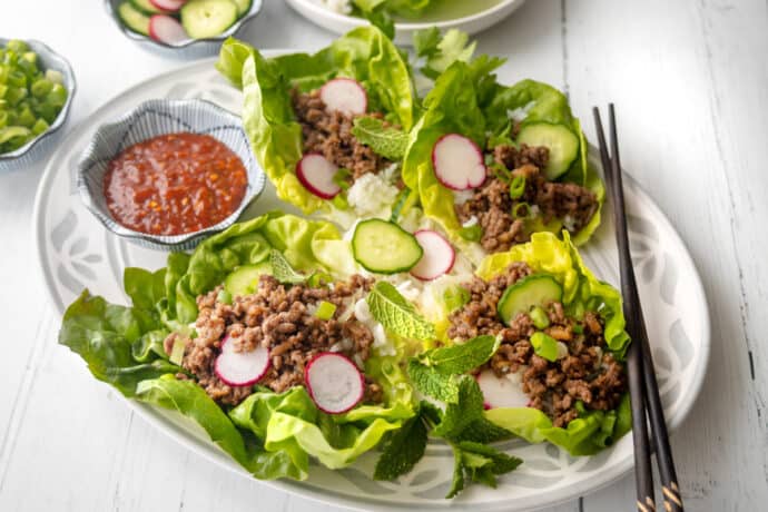 A plate of lettuce wraps filled with Korean ground beef, sliced radishes, cucumber, green onions, and mint leaves, served with a bowl of dipping sauce and chopsticks on a white table.