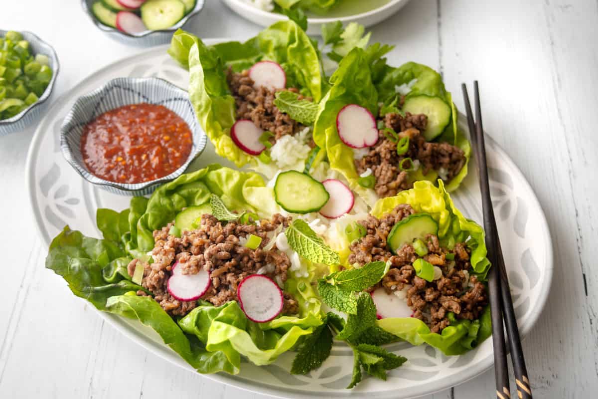 A plate of lettuce wraps filled with Korean ground beef, sliced radishes, cucumber, green onions, and mint leaves, served with a bowl of dipping sauce and chopsticks on a white table.