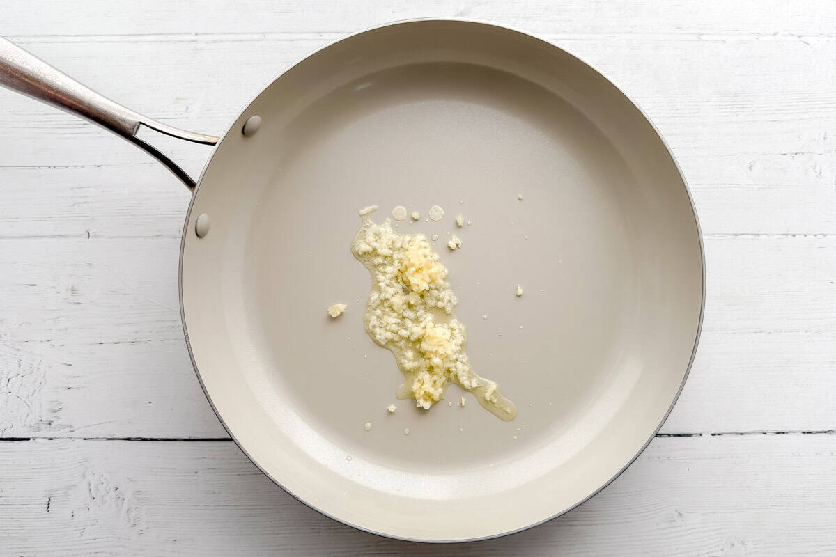A stainless steel frying pan with minced garlic and oil sits on a white wooden surface.