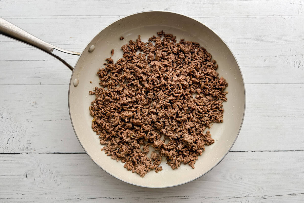 Cooked ground beef in a large light-colored frying pan sits on a white wooden surface, ready to be used in a recipe.
