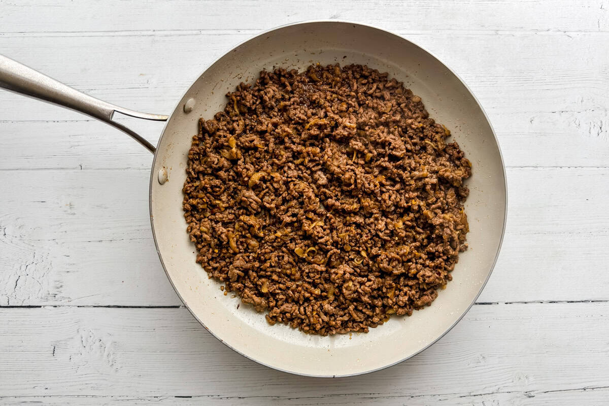 A frying pan filled with cooked and browned ground beef sits on a white wooden surface.