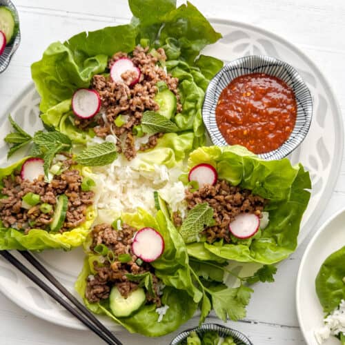 A plate of lettuce wraps filled with ground meat, radish slices, cucumber, mint leaves, and a side of rice. A bowl of red dipping sauce and more rice are nearby, with chopsticks on the side.