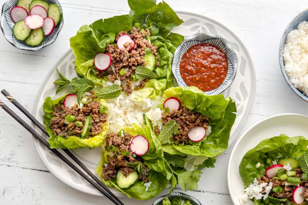 A plate of lettuce wraps filled with ground meat, radish slices, cucumber, mint leaves, and a side of rice. A bowl of red dipping sauce and more rice are nearby, with chopsticks on the side.