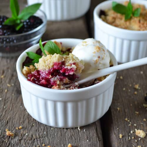 A small white ramekin filled with berry crumble and a scoop of vanilla ice cream, garnished with a mint leaf, sits on a rustic wooden surface with blackberries and crumbs scattered around.