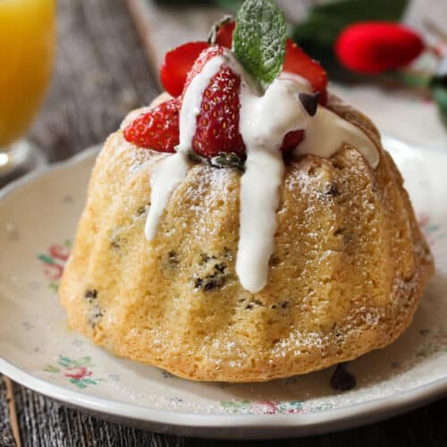 A small bundt cake topped with whipped cream, fresh strawberries, and a mint leaf sits on a floral-patterned plate, with a glass of orange juice and a red rose in the background on a rustic wooden table.