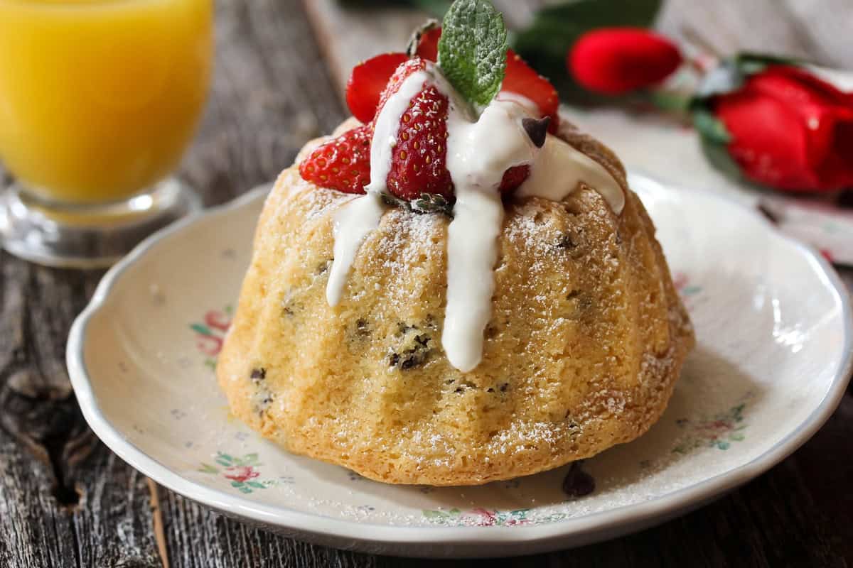 A small bundt cake topped with whipped cream, fresh strawberries, and a mint leaf sits on a floral-patterned plate, with a glass of orange juice and a red rose in the background on a rustic wooden table.