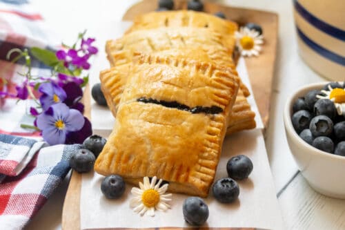 Golden-brown blueberry hand pies filled with sweet jam are stacked on parchment paper, surrounded by fresh blueberries, small daisies, and purple flowers on a wooden board near a bowl of blueberries.