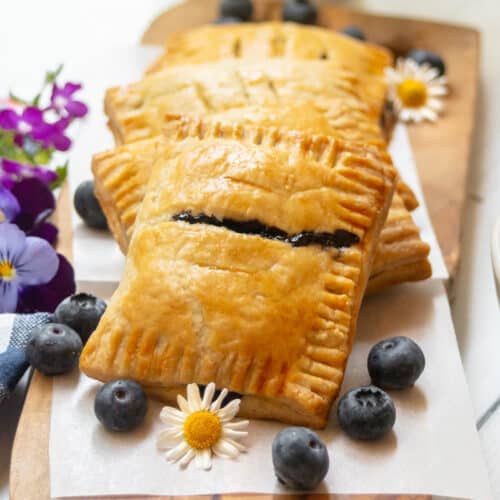 Golden-brown blueberry hand pies filled with sweet jam are stacked on parchment paper, surrounded by fresh blueberries, small daisies, and purple flowers on a wooden board near a bowl of blueberries.