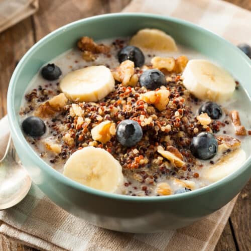 A bowl of quinoa porridge topped with banana slices, blueberries, and walnuts, served with milk. The bowl is on a wooden table with a checkered cloth and a spoon beside it.