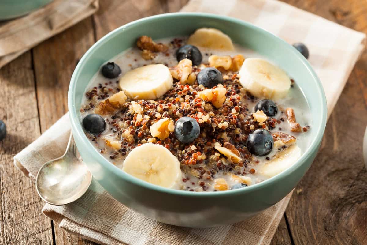 A bowl of quinoa porridge topped with banana slices, blueberries, and walnuts, served with milk. The bowl is on a wooden table with a checkered cloth and a spoon beside it.