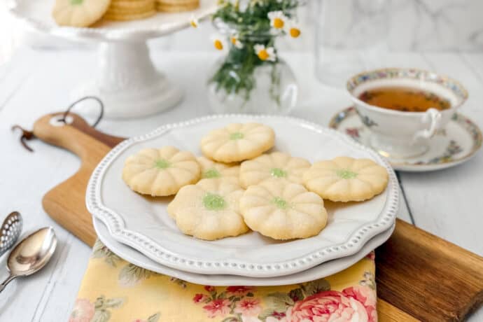 A white plate holds six flower-shaped sugar cookies with green centers, reminiscent of Girl Scout Cookies, placed on a floral napkin. Nearby are a teacup, saucer, spoons, and a vase of small white flowers on a wooden board.
