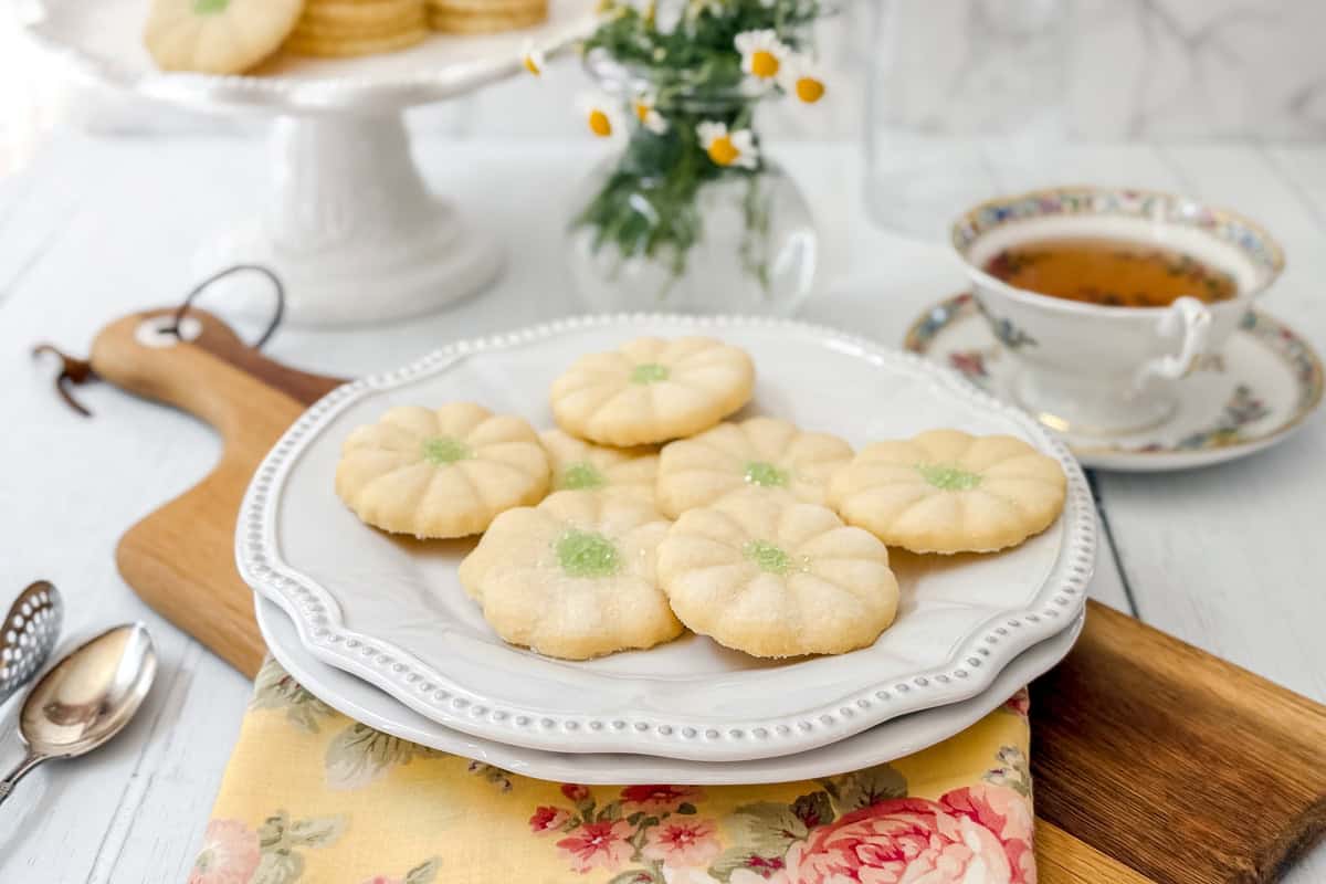 A white plate holds six flower-shaped sugar cookies with green centers, reminiscent of Girl Scout Cookies, placed on a floral napkin. Nearby are a teacup, saucer, spoons, and a vase of small white flowers on a wooden board.