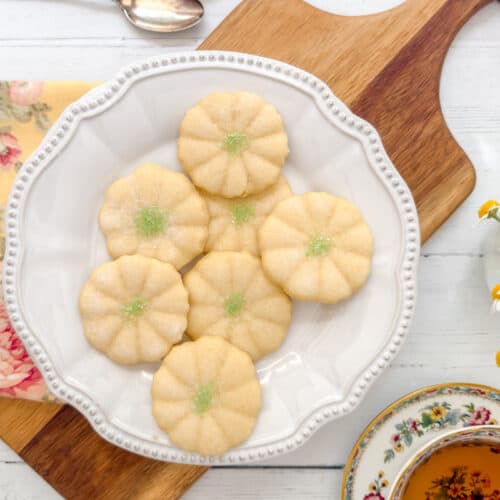 A white plate with seven flower-shaped cookies, reminiscent of Girl Scout Cookies and sprinkled with green sugar, sits on a wooden board. A floral napkin, a cup of tea, and small daisies are nearby on a white wooden table.