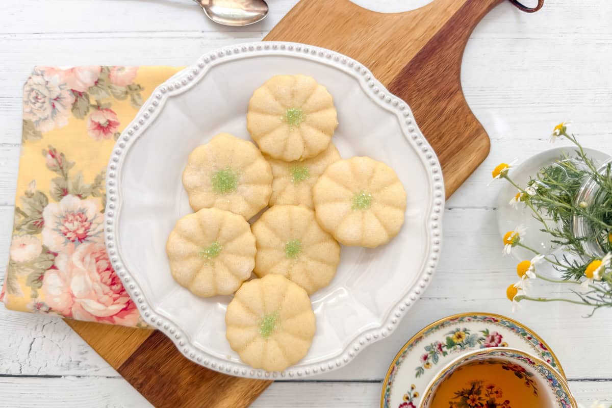 A white plate with seven flower-shaped cookies, reminiscent of Girl Scout Cookies and sprinkled with green sugar, sits on a wooden board. A floral napkin, a cup of tea, and small daisies are nearby on a white wooden table.